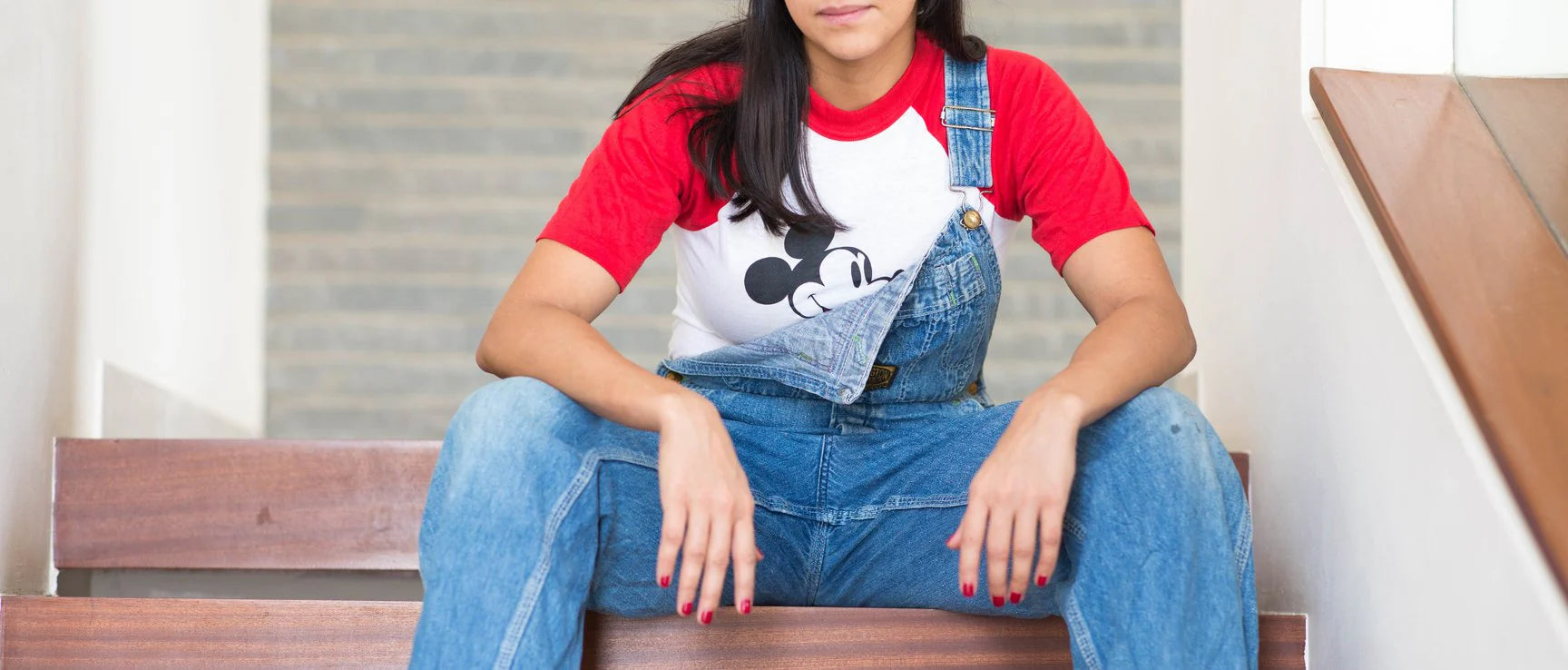 Person wearing a red shirt with a graphic design and blue overalls sitting on a bench.